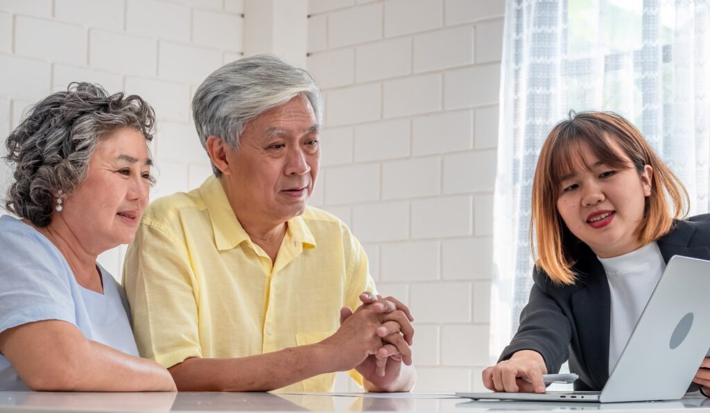 Asian older adult couple seated at a table getting advice from a younger Asian woman as they look at a laptop