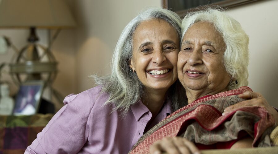 a grey-haired woman of color and her older mother smile at camera while together on couch