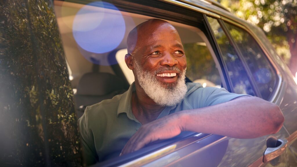African-American man with a white beard smiles widely as he sits in the back seat of a moving car, with his arm resting on the open window