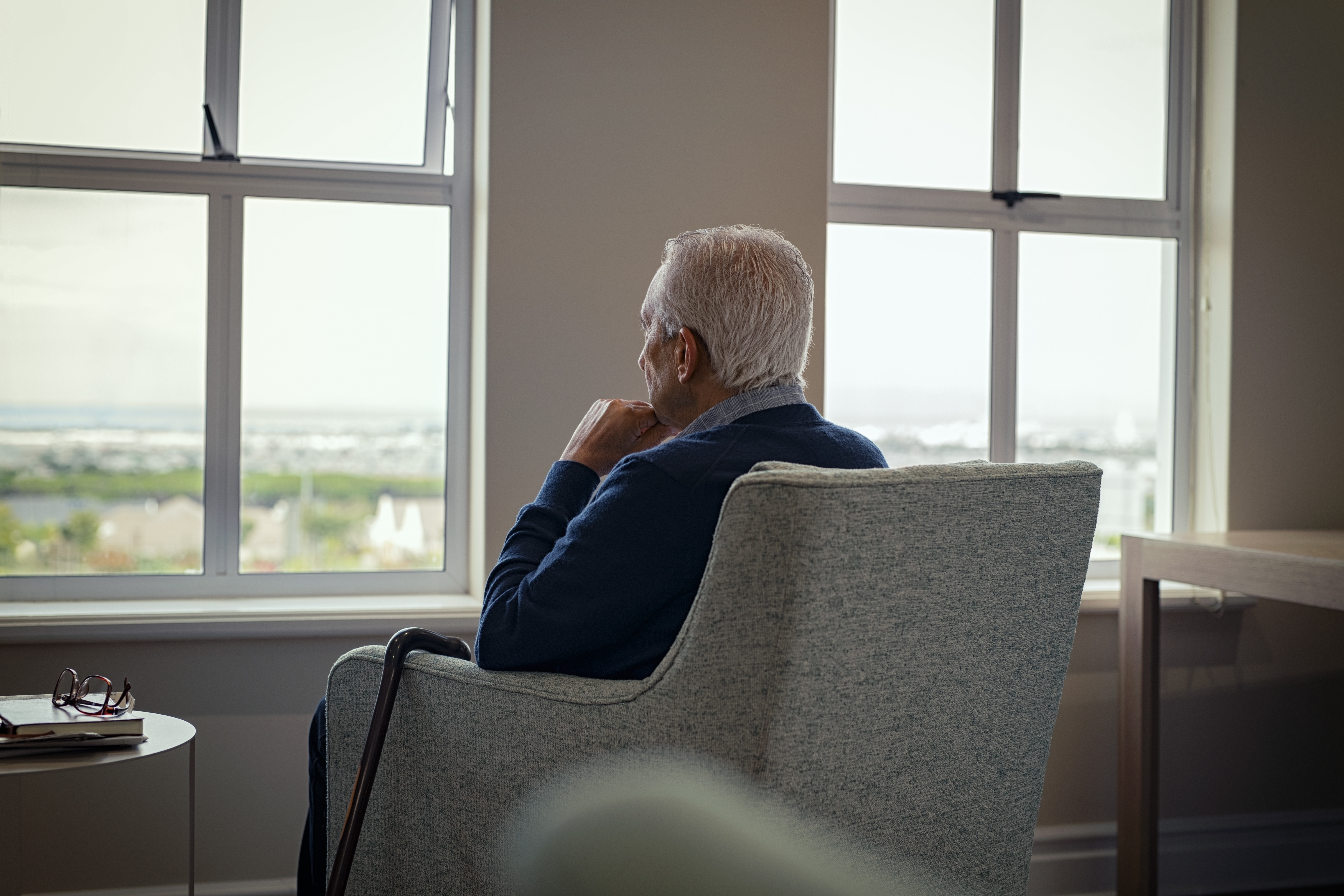 Photo of a man seated in an armchair in a nursing facility, seen from the back as he looks out the window