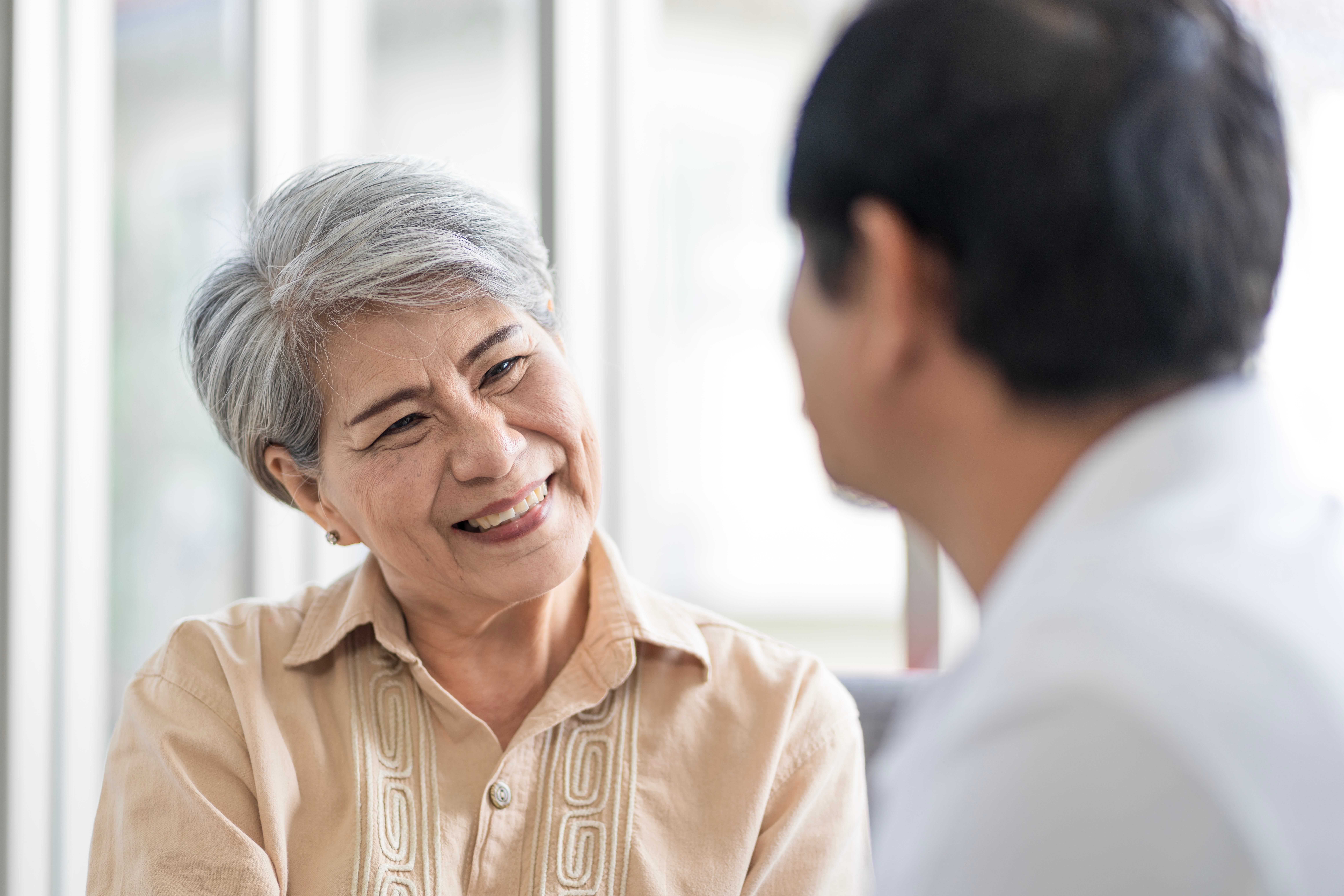 Older Asian woman with short white hair smiles as she sits and speaks to a dark-haired man. He seen from behind, face not shown, and slightly out of focus.