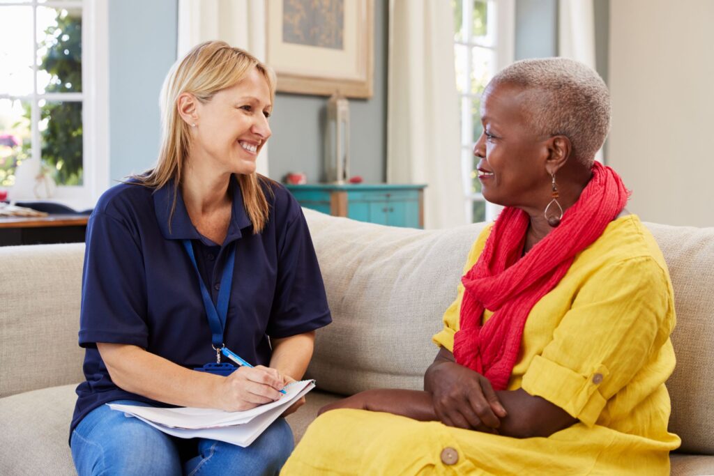 Female worker with blonde hair and a navy blue shirt smiles as she takes notes while talking to a client on the client's sofa. The client is an older woman of color with white hair clipped very close to the head, wearing a bright yellow dress and bright orange scarf, and smiling at the worker