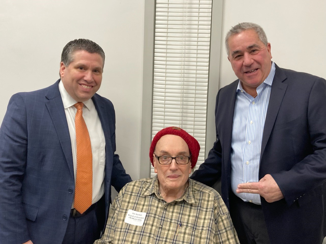 Two MA lawmakers, wearing suits and ties, stand with Jon Norton of the MVES Board seated between them. Jon is wearing one of his trademark hats, which is red. MA Sen. Sal DiDomenico is at left. MA Rep. Joe McGonagle is gesturing at Mr. Norton with two fingers.