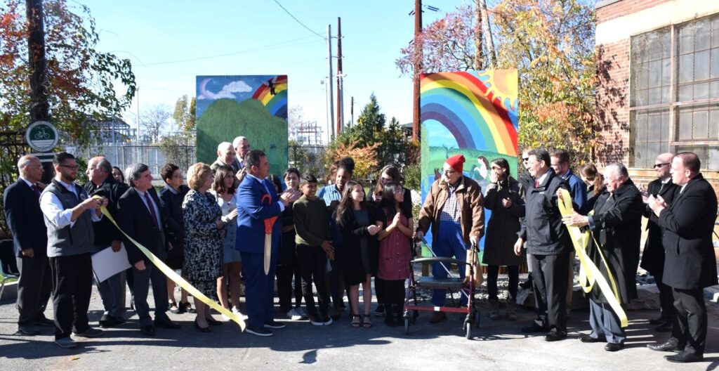 Large group of students and city officials lined up in a row, the ribbon has just been cut on the project, and everyone is clapping as they turn toward Jon Norton. In the background is a large two-piece painting by students showing the outdoors, rainbows, sub, etc.