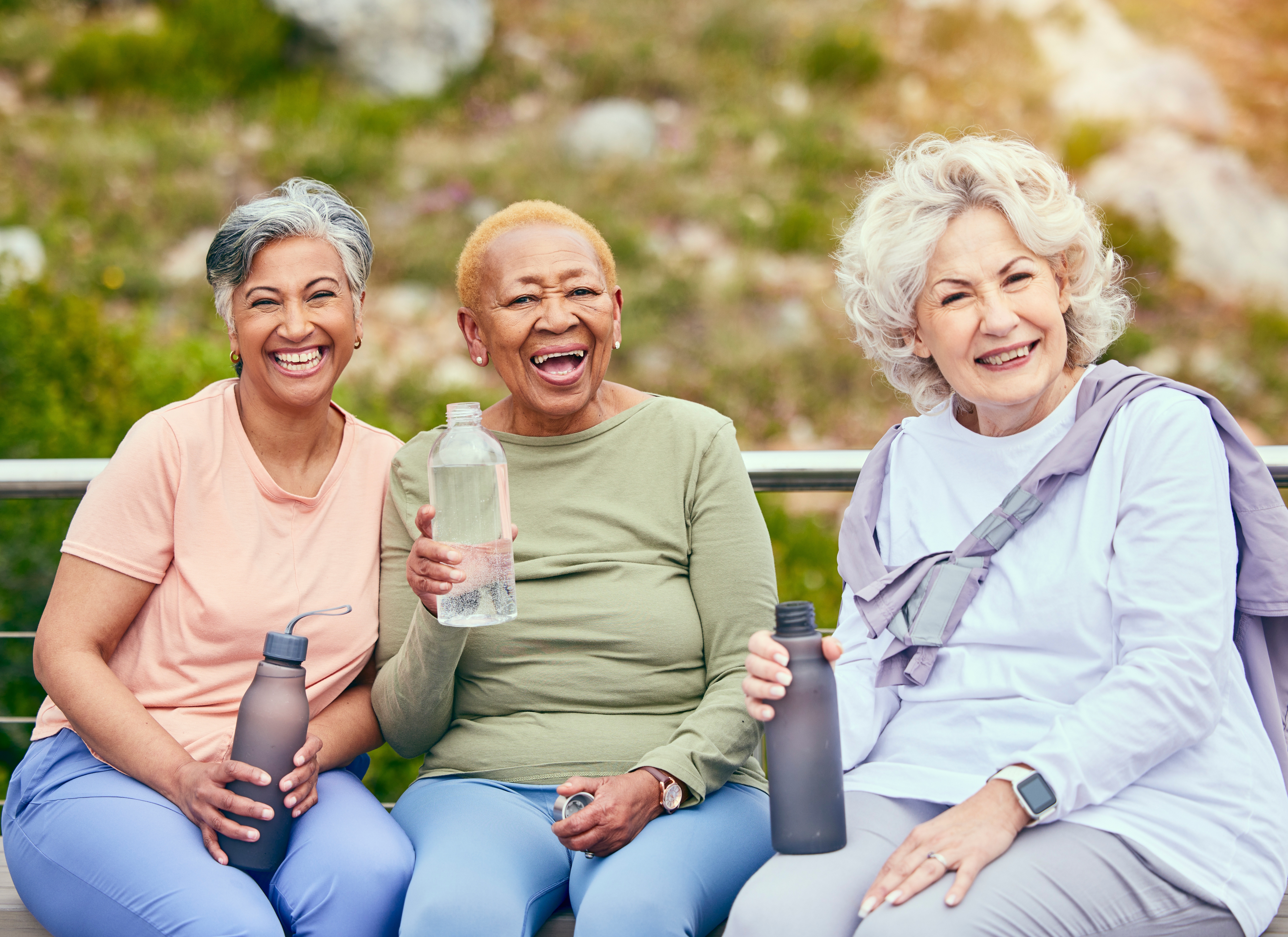 Three older adult women seated on a bench in a park. They are dressed in casual exercise type clothing, holding water bottles, and smiling as they look into the camera.