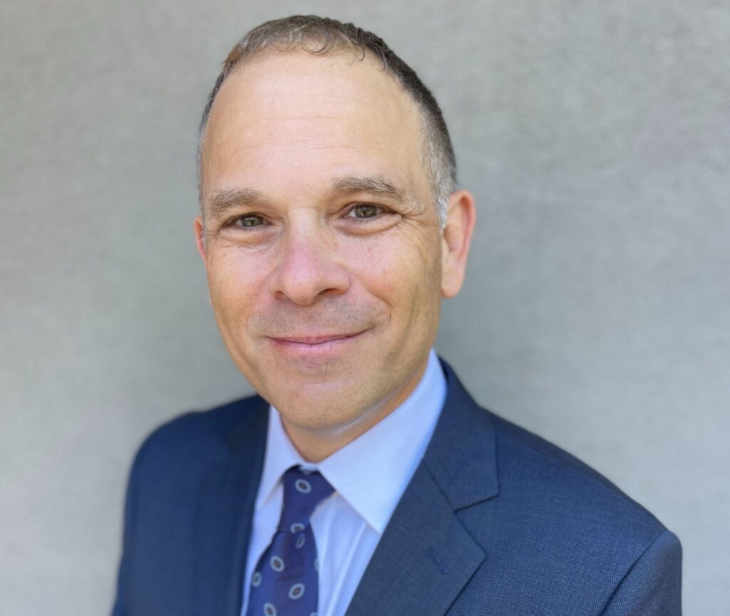 Dr. Edward Alan Miller shown from the shoulders up, against a grey background. He is smiling and looking directly into the camera. Wearing a dark blue suit, bright blue necktie printed with small circular shapes, and a light blue dress shirt.