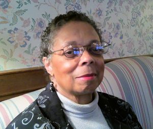 Mae Upperman, with short grey and black hair, wearing wire rimmed glasses and a black printed blazer, seated on a couch.