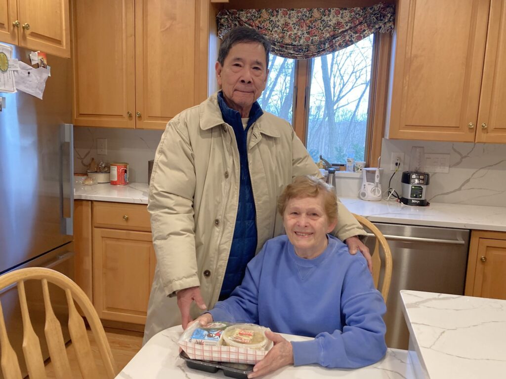 Brenda Kerrigan, an older adult woman, who is seated at her kitchen table with the meal that's just been delivered. Frank Liu, an MVES driver, stands with his hand on her shoulder.