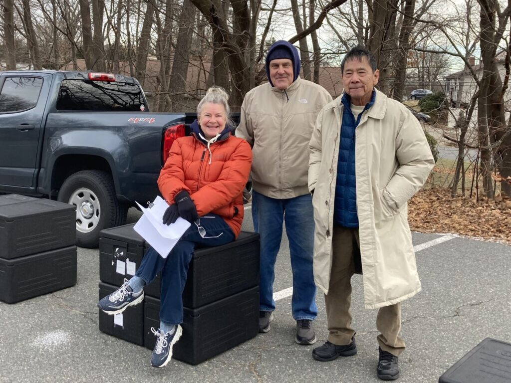 A woman and two men, bundled up in winter clothing, smile at the camera in a parking lot surrounded by trees. There are large black containers which contain meals for delivery, and the woman is seated on one of these, while the men stand next to her.