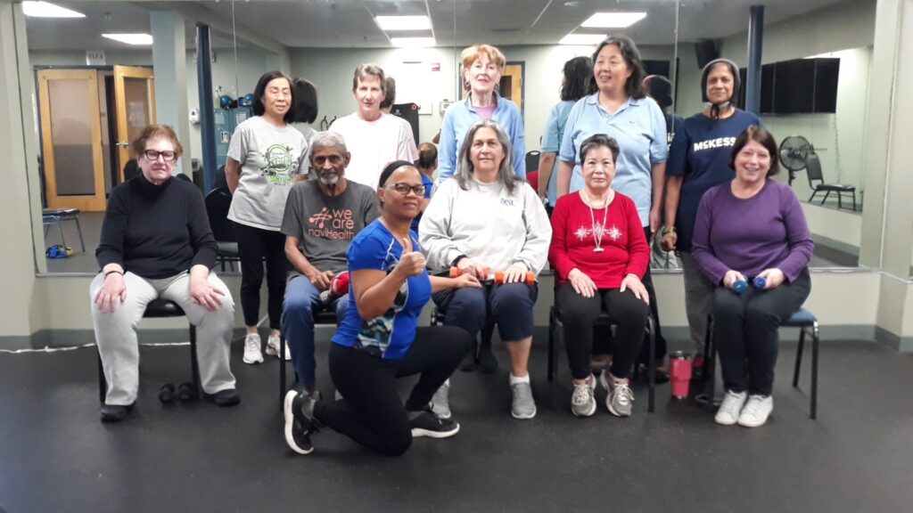 10 older adults wearing athletic clothing appear in front of a mirror in a fitness room. Some are standing and some are seated. Their instructor is kneeling in front, giving a thumbs up.