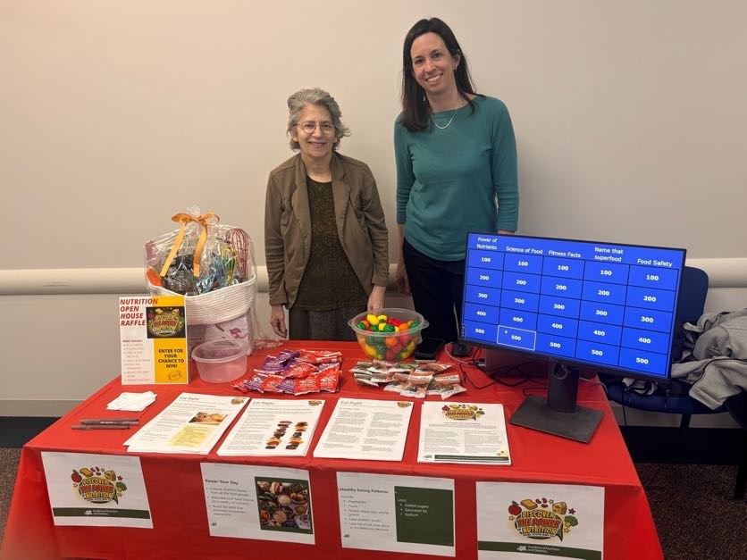 Two staff members stand at table with nutritional info, packets of healthy snacks and monitor displaying a food quiz.