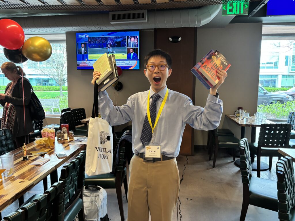 College student who volunteers is displaying books in each hand that he won in raffle. He has a surprised happy expression.